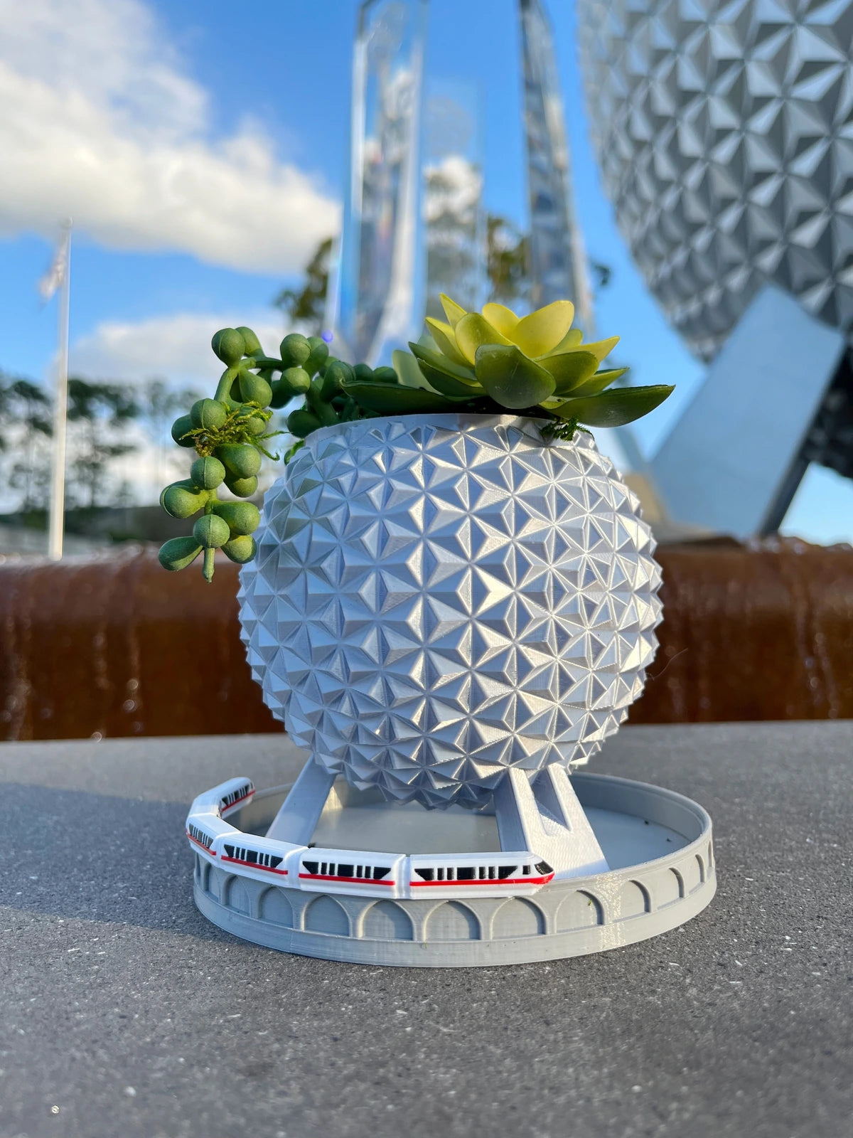 Silver Grand and Miraculous Planter with succulents and a Monorail Drip Tray in front of Spaceship Earth